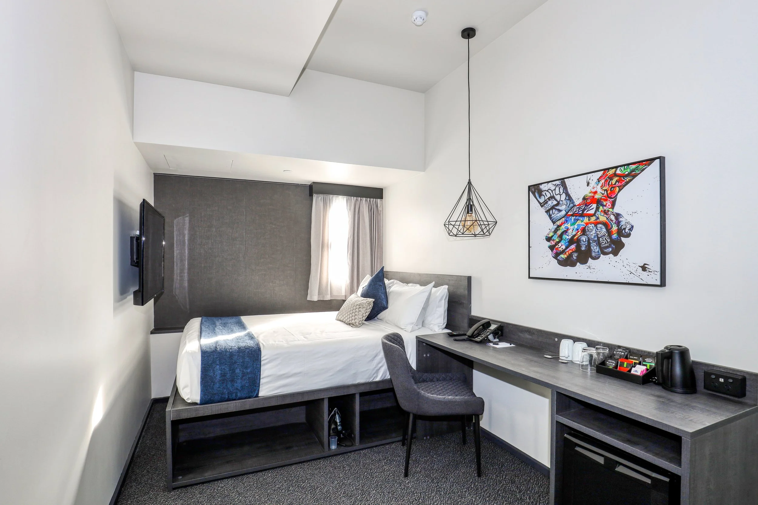 The Viv Hotel Queen Room with a queen bed under a small window letting in soft natural light